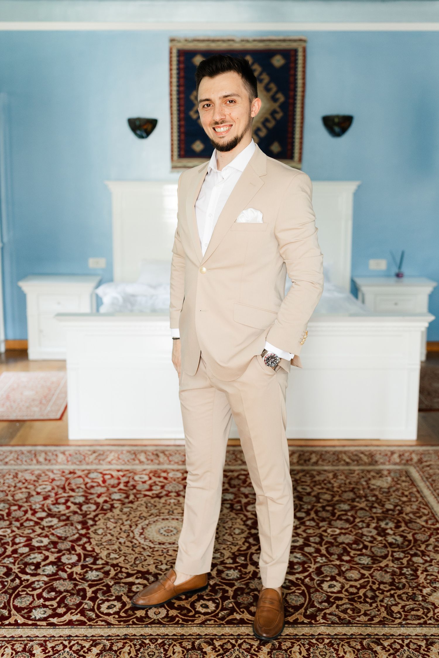 A person in a beige suit and brown shoes stands on an ornate rug in an elegant room with blue walls.