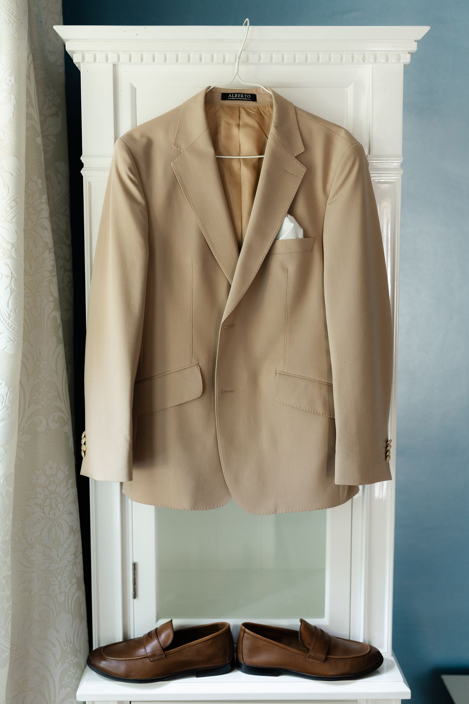 Beige blazer jacket displayed on white wall-mounted hooks against a blue-gray wall.
