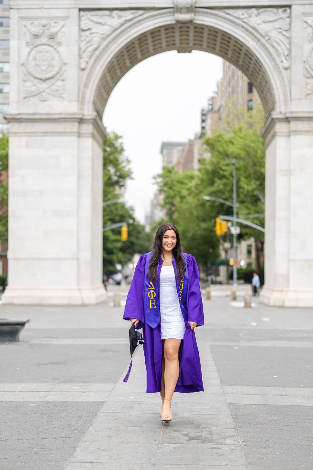 Graduate poses under Washington Square Arch in purple graduation regalia and white dress.