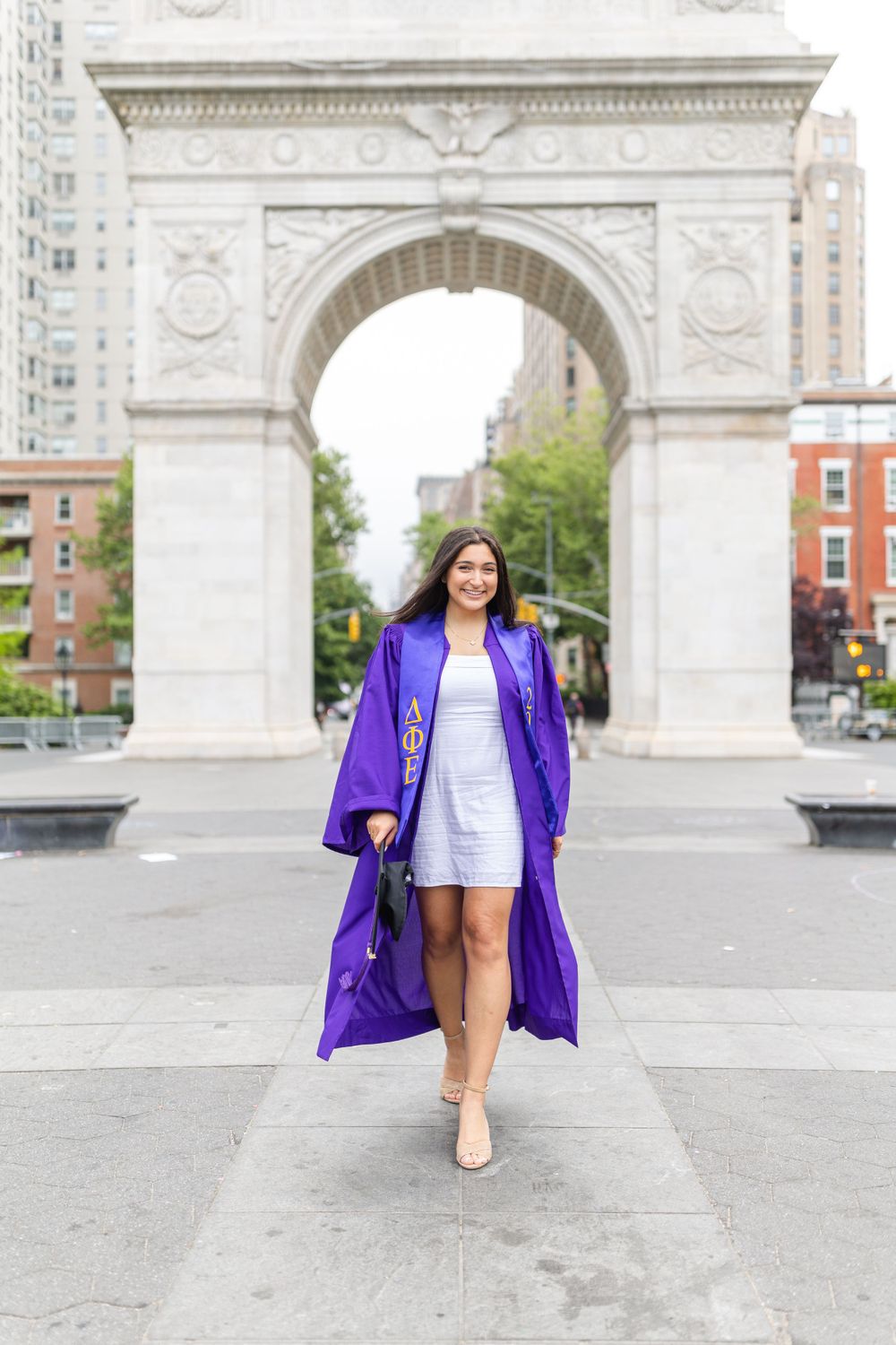 Graduate strides through Washington Square Park wearing purple graduation gown and white dress.