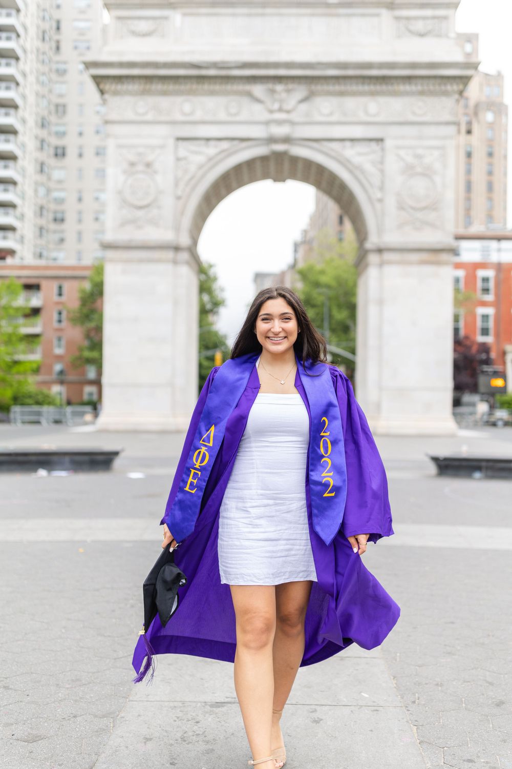 Graduate smiles in purple graduation gown and white dress at Washington Square Park.