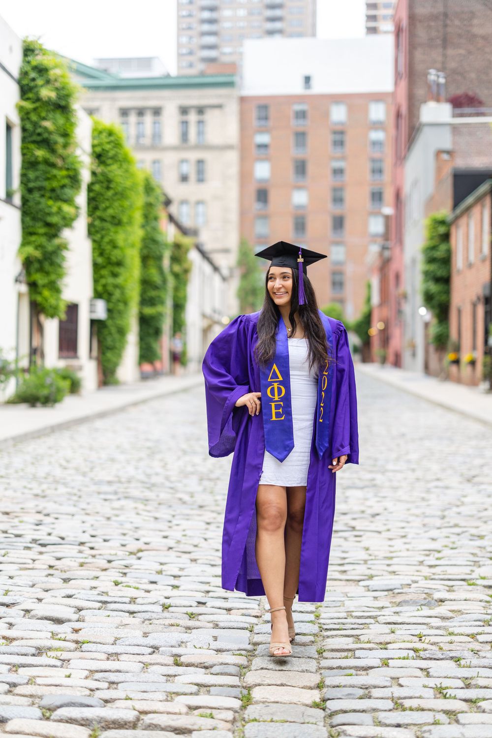 Graduate walking on cobblestone street in purple graduation gown and cap.