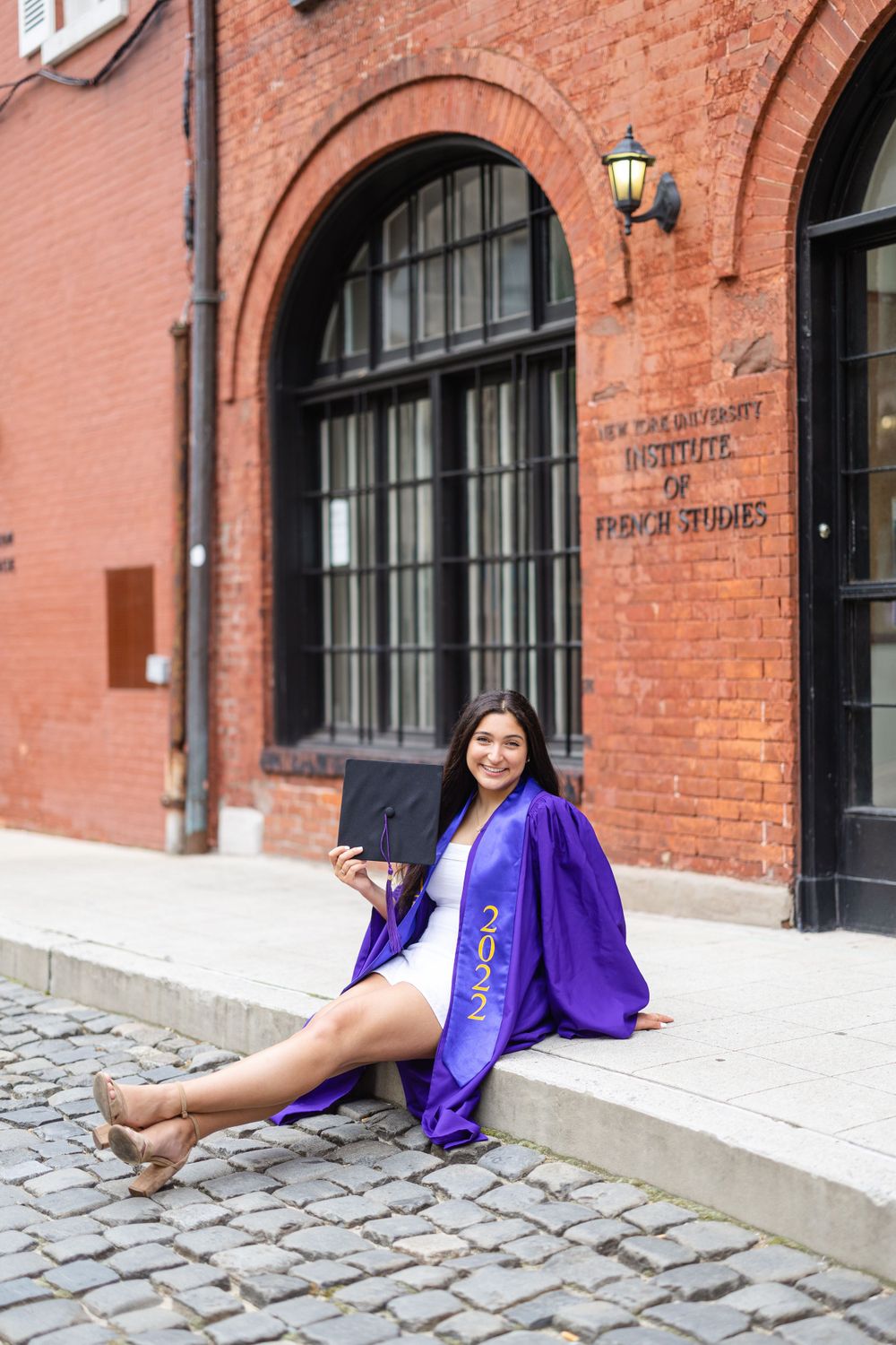 Graduate sitting on steps of brick building in purple graduation gown and white dress.