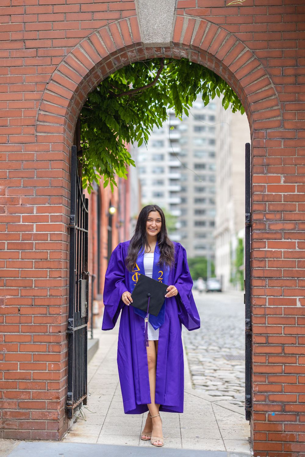 Graduate in purple NYU commencement robe poses under brick archway entrance.
