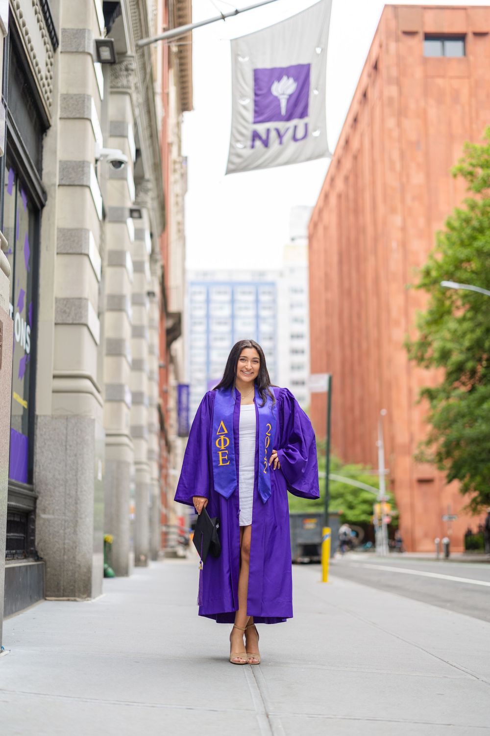 Graduate in purple regalia walks on campus with NYU flag in background.