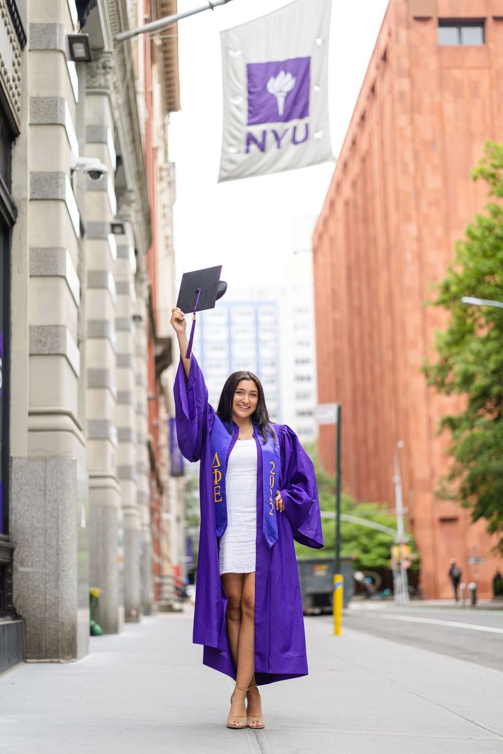Graduate throws cap in air while wearing purple NYU graduation gown on campus street.