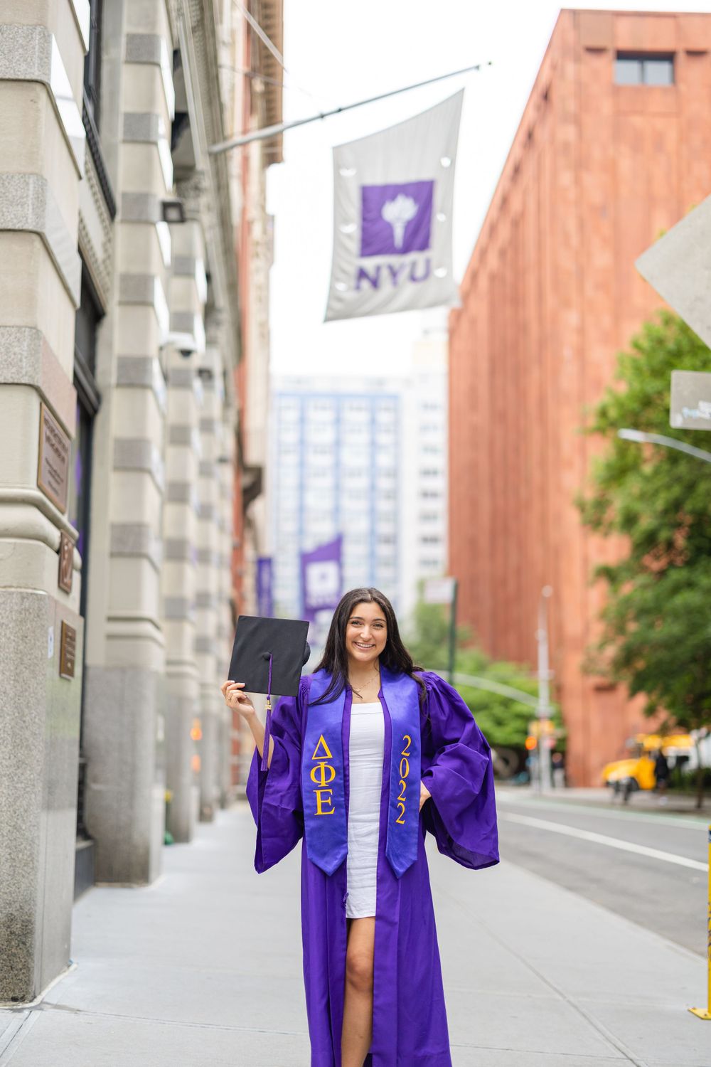 Graduate holds diploma while standing beneath NYU flag on campus.