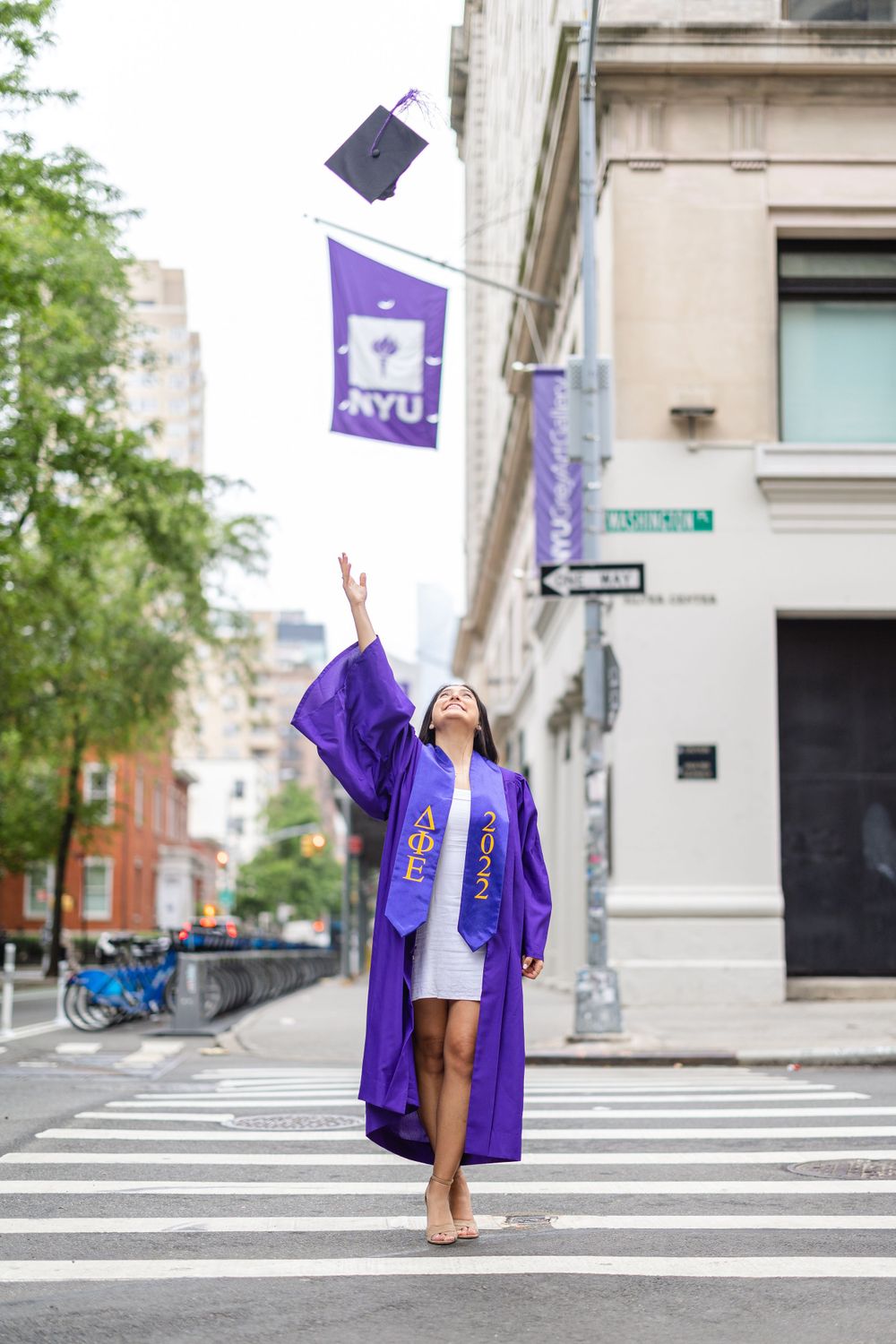 Graduate tosses graduation cap in the air at intersection with NYU flag visible.