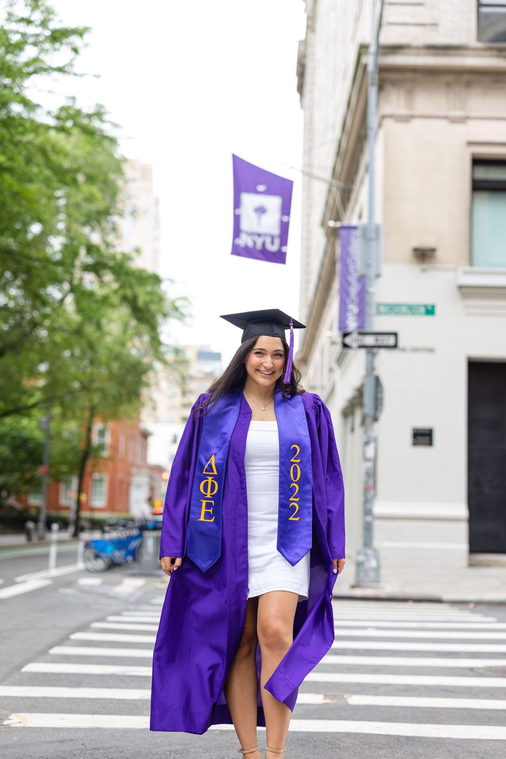 Graduate poses in traditional purple NYU regalia on city street.