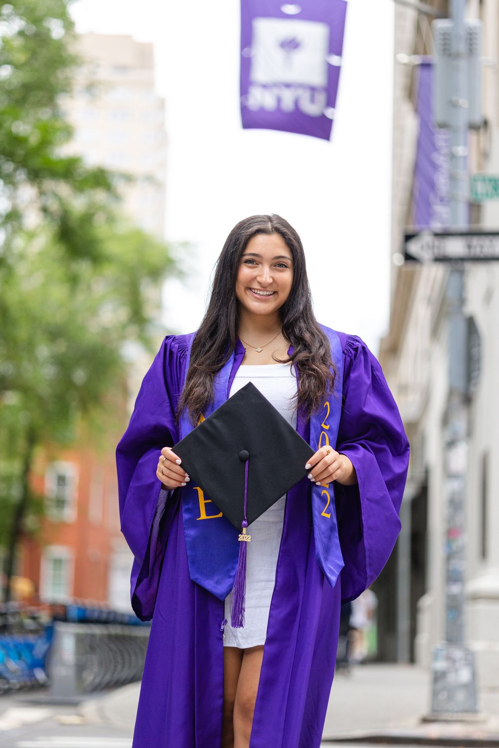 Graduate displays graduation cap while wearing purple NYU commencement robe outdoors.