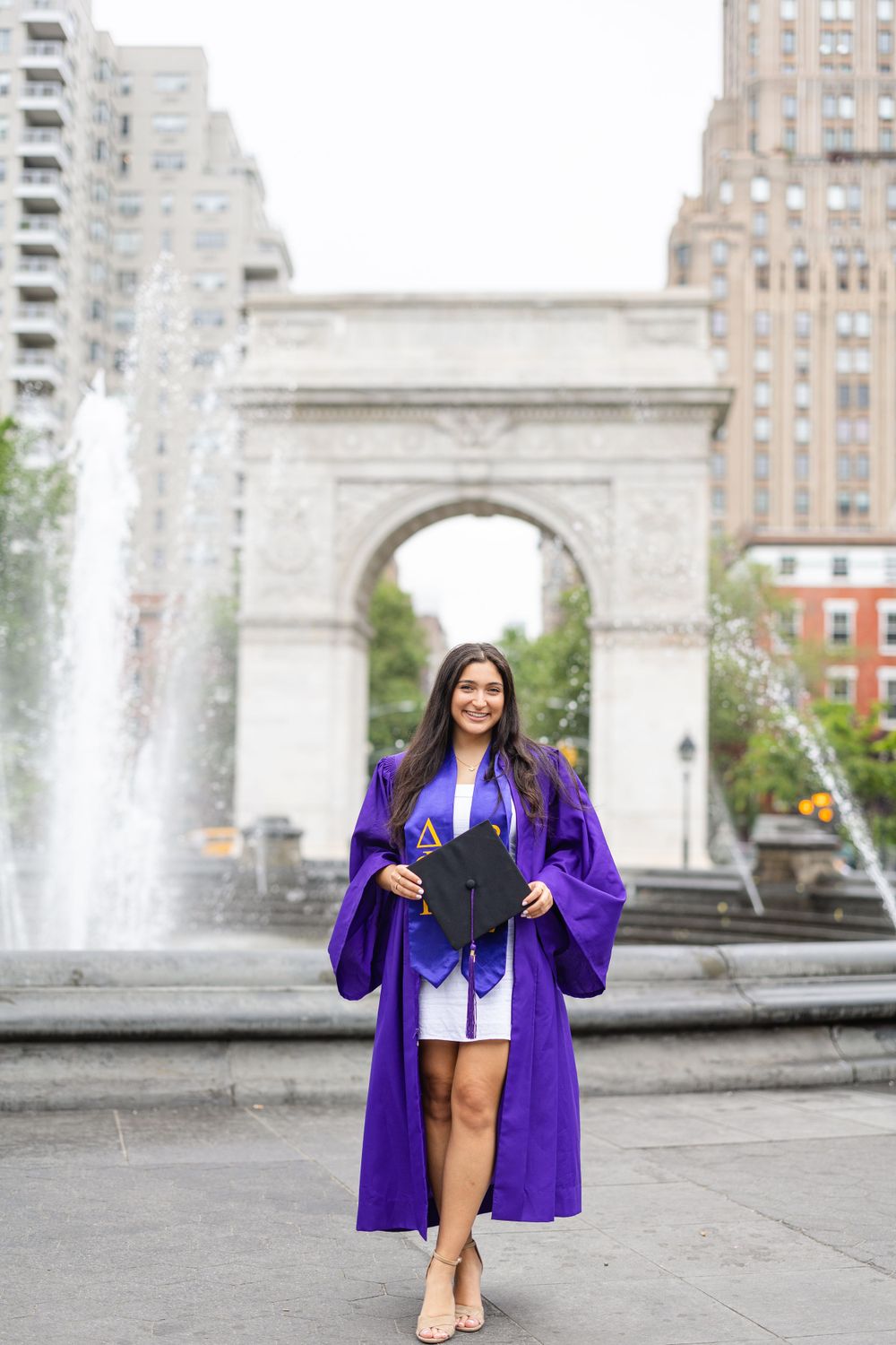 Graduate poses with diploma in front of Washington Square Arch with fountains.