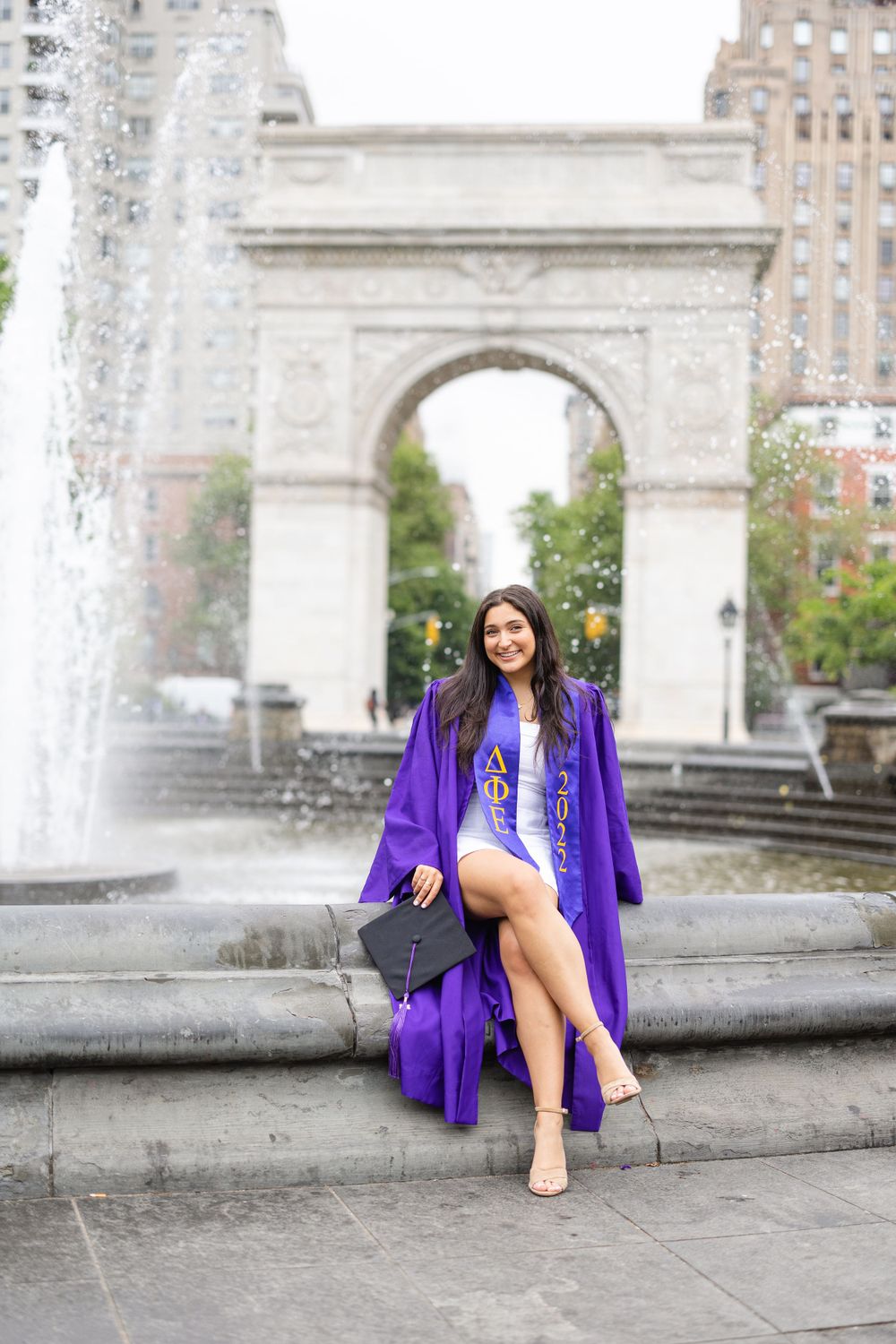 Graduate poses on fountain steps with Washington Square Arch in background.