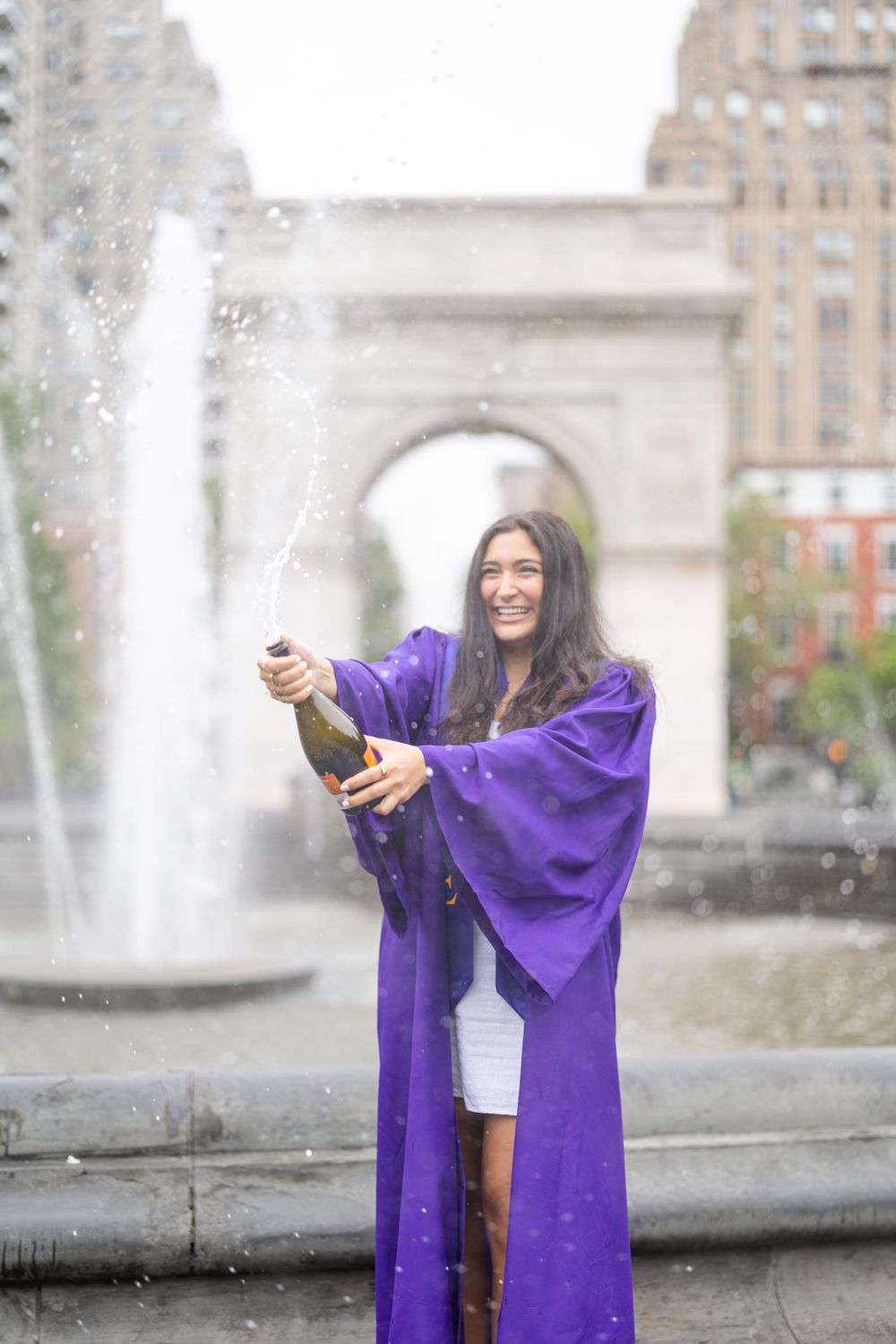 Graduate celebrates with champagne by fountain at Washington Square Park.