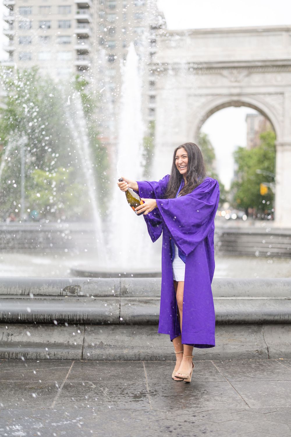 Graduate in purple graduation gown celebrates with champagne by fountain at Washington Square Park.