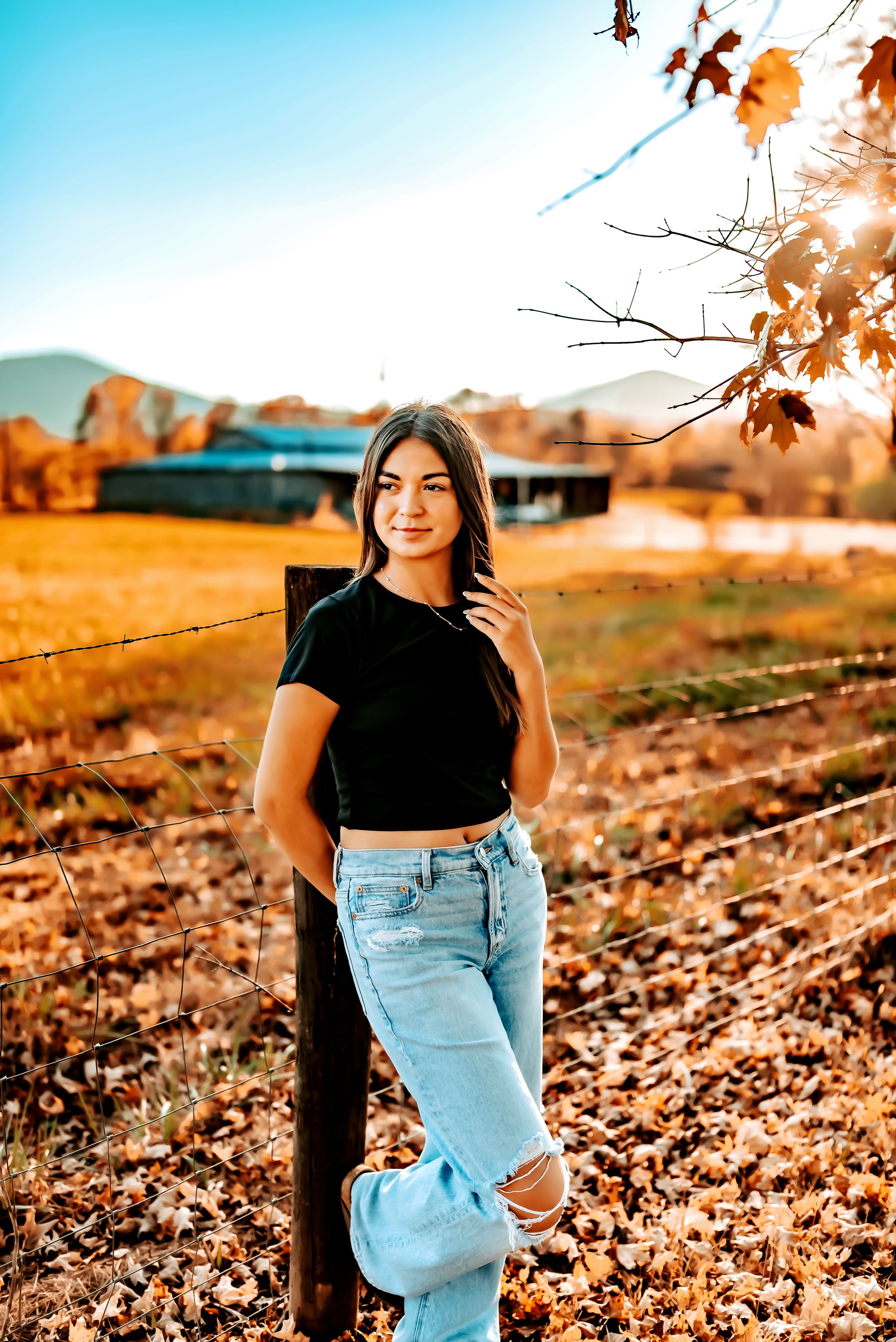A person in a black top and jeans poses against an autumn backdrop with golden leaves and warm sunlight.