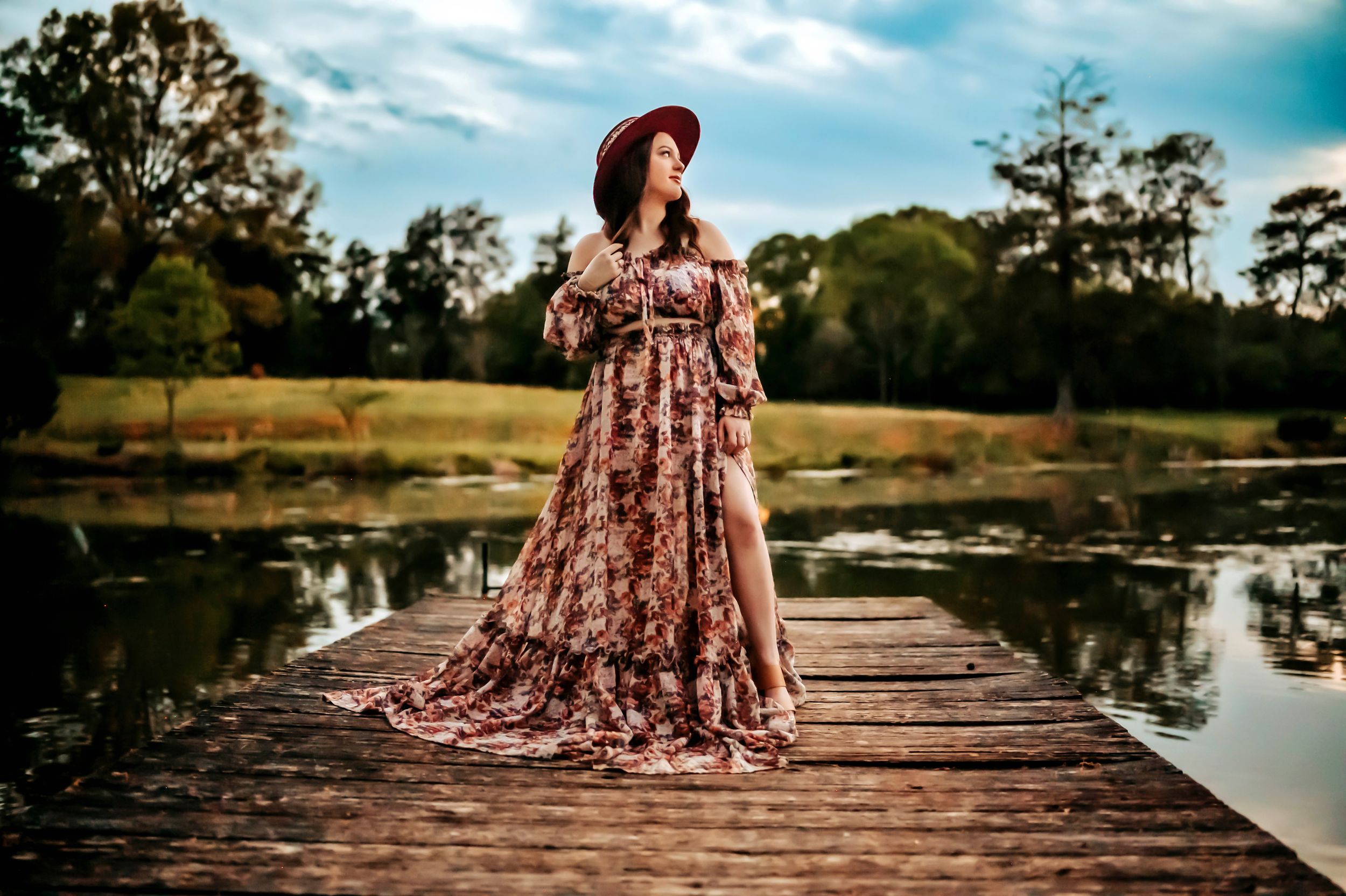 Woman in floral maxi dress and wide-brim hat poses on wooden dock by serene lake surrounded by lush trees.