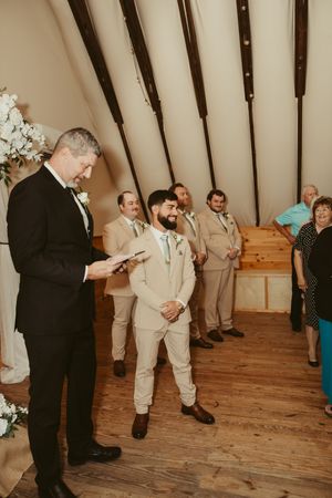 Groom in beige suit smiling at altar with groomsmen and officiant in rustic barn chapel.