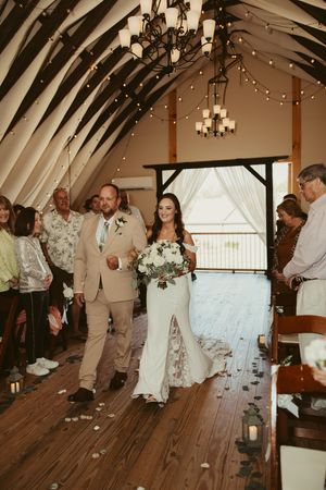 Bride walking down aisle in white gown with bouquet beside escort in barn wedding venue.
