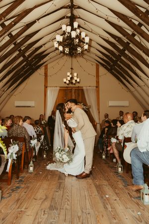 Newlyweds sharing first kiss in barn aisle surrounded by guests and chandelier lighting.