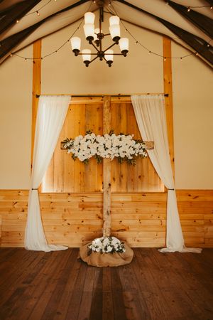 Rustic barn wedding altar with wooden cross, white floral arrangements, draped fabric, and chandelier.