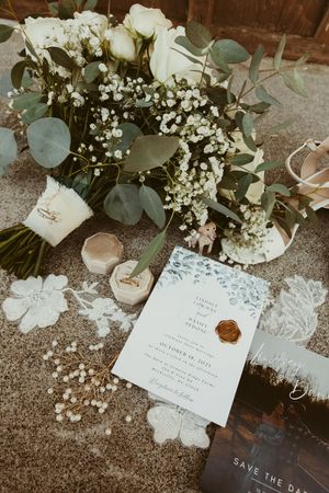 Wedding flat lay with white rose bouquet, eucalyptus, ring boxes, lace, and floral invitation on sand.