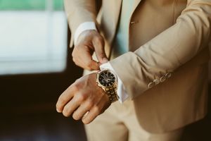 Groom in beige suit adjusting gold watch and white dress shirt cufflink on wedding day.
