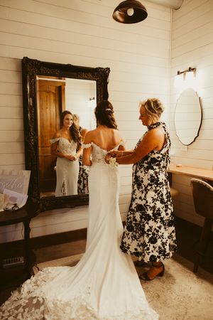 Mother helping bride button up elegant white lace wedding dress in rustic bridal suite.