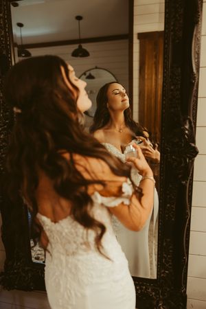 Bride in lace wedding gown applying perfume while gazing into ornate mirror before ceremony.