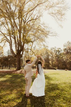 Groom twirls bride in white gown and veil on green grass beneath a large tree outdoors at The Barn at Granite Ridge Farm in Mocksville, NC.