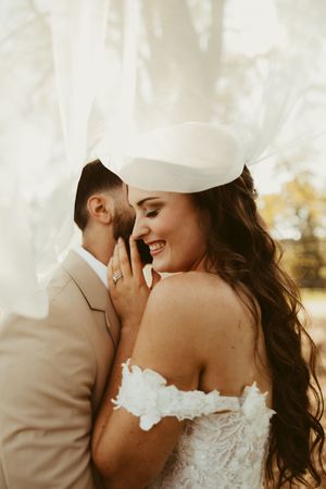 Smiling bride in off-shoulder lace gown and white hat leans into groom during outdoor wedding portraits.