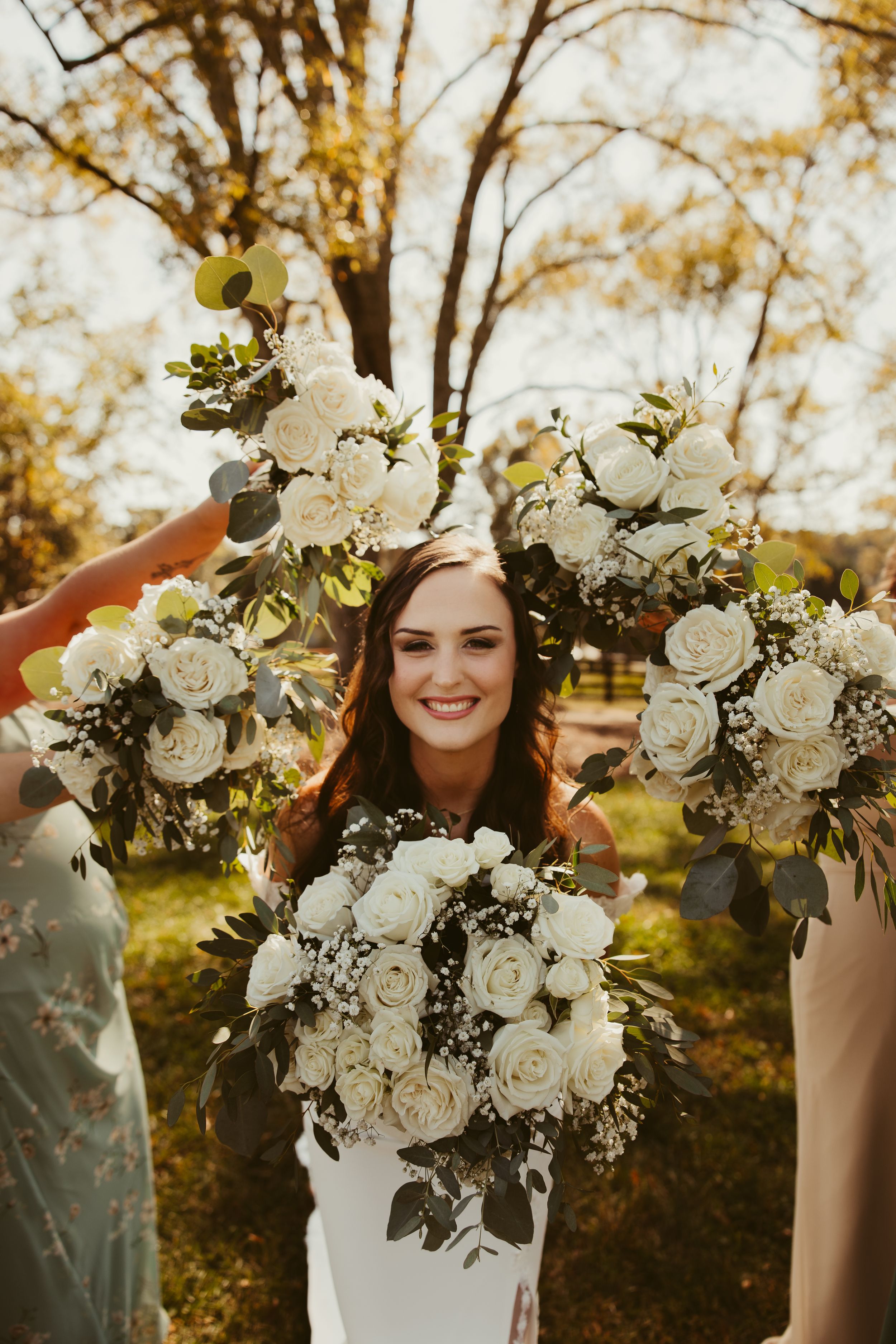 Smiling bride surrounded by white rose and eucalyptus bouquets held up by bridesmaids outdoors.