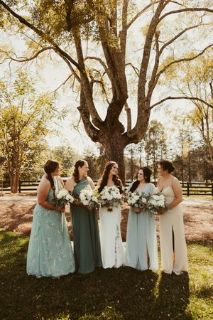 Bride with bridesmaids in sage and mint gowns holding white bouquets under large oak tree.