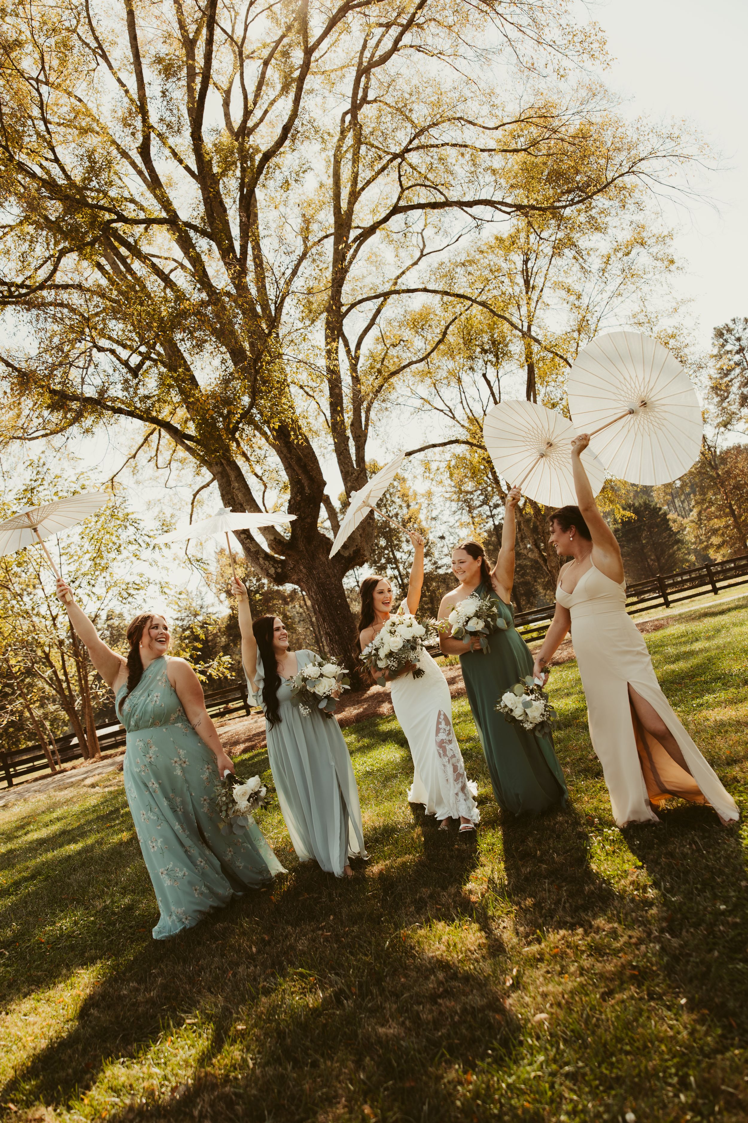 Bridesmaids in green dresses raise white parasols outdoors under a large tree on a sunny wedding day.