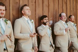 Groomsmen in tan suits smoke cigars while leaning against a rustic wooden barn wall.