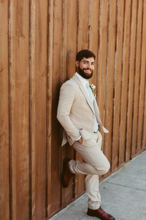 Smiling groom in a beige suit with boutonniere leans against a wooden wall on his wedding day.