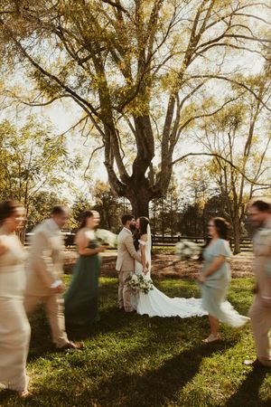 Bride and groom share a kiss under a large tree as blurred wedding party members walk past.