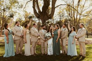 Wedding party poses outdoors in sage green and tan attire beneath a large tree at The Barn at Granite Ridge Farm.
