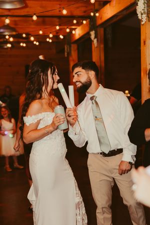 Bride and groom dance joyfully holding glow sticks at their rustic barn wedding reception with string lights overhead.
