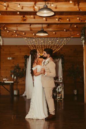 Bride and groom share first dance under string lights in a warmly lit rustic barn venue.