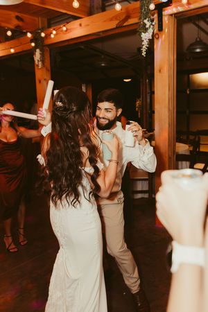 Smiling groom dances with bride holding glow sticks at a warmly lit rustic barn wedding reception.