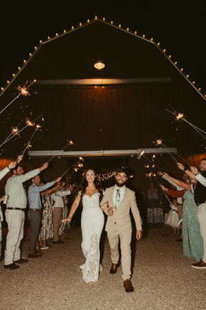 Bride and groom exit through a sparkler tunnel outside a barn venue at their nighttime wedding reception.