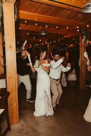 Bride and groom dance together with glow sticks among guests at a rustic barn wedding reception.
