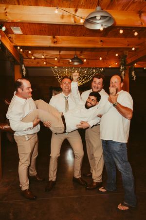 Groom and groomsmen laugh and pose playfully together under string lights at a rustic wedding reception.