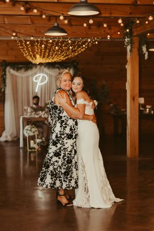Bride in lace gown embraces her mother on the dance floor of a rustic barn wedding reception.