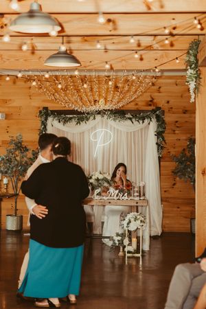 Guest couple dances as bride watches from sweetheart table in a decorated rustic barn reception hall.