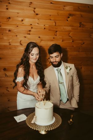 Bride and groom cut their elegant single-tier white wedding cake against a warm wooden wall backdrop.