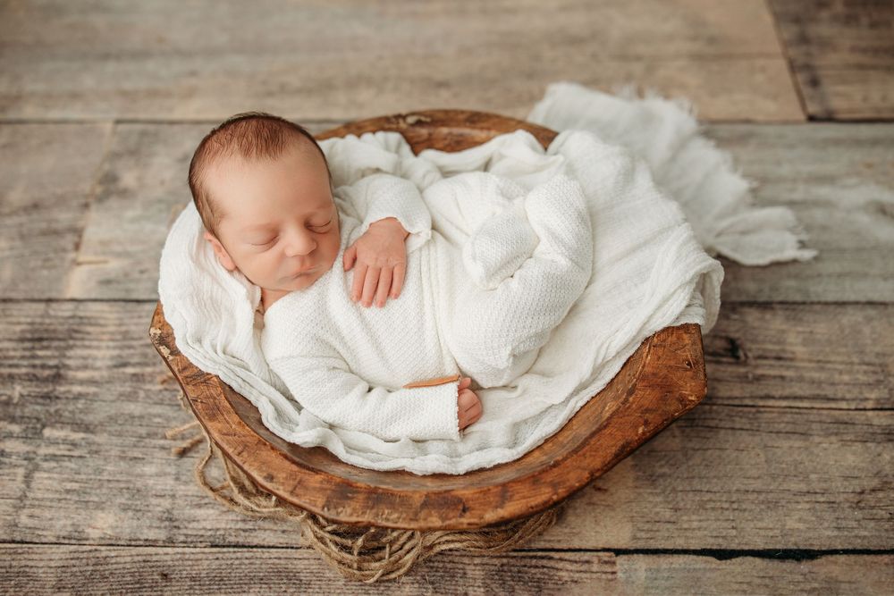 A sleeping newborn wrapped in white fabric rests peacefully in a rustic wooden basket on wooden floor.