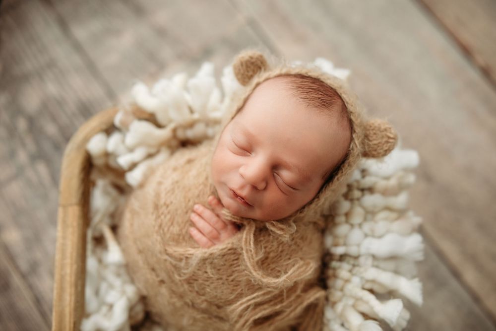 Sleeping newborn baby wearing cozy knit bear hat rests peacefully on wooden backdrop.