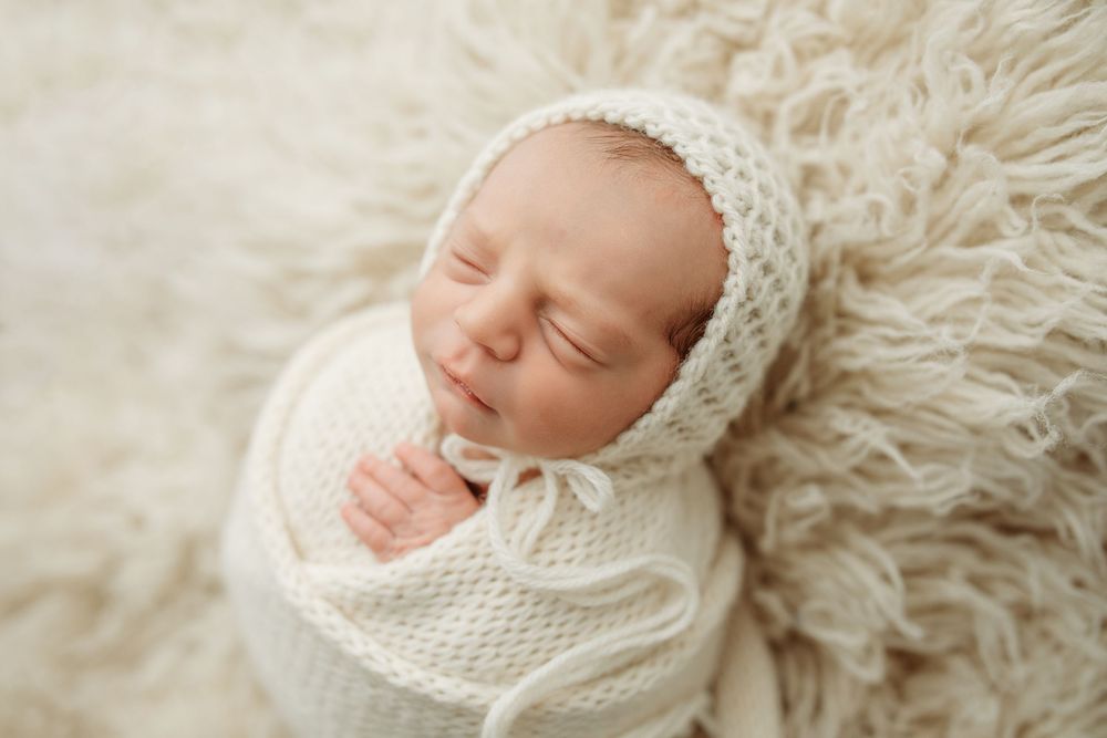 Peaceful sleeping newborn wrapped in soft white knit blanket and bonnet on fluffy cream backdrop.