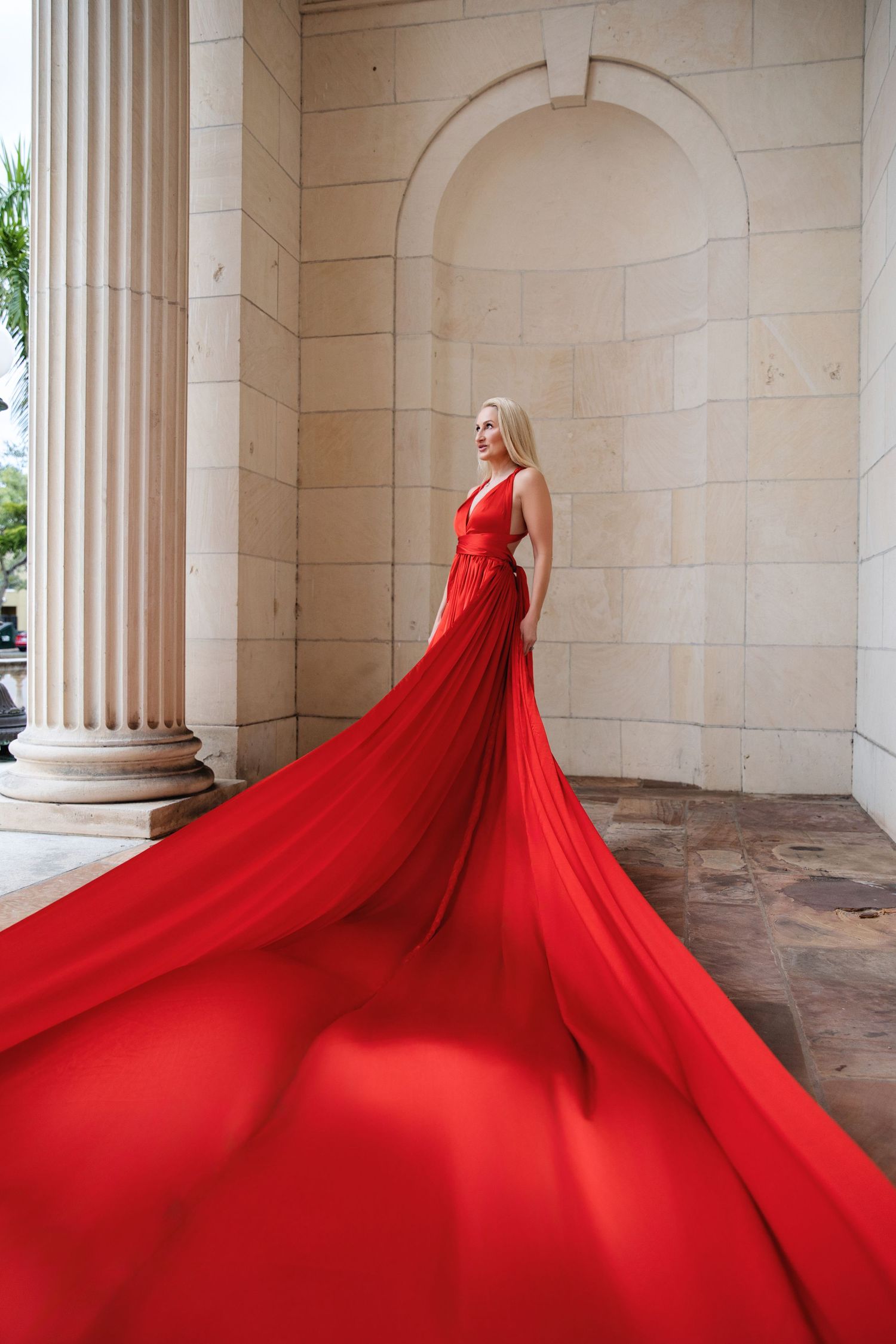 A flowing red gown with dramatic cathedral-length train cascades across marble columns in an elegant architectural setting.